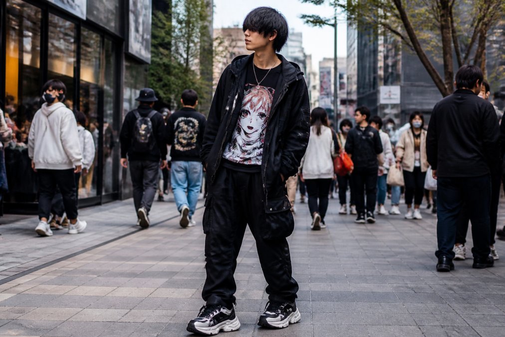 A stylish young adult on a busy city sidewalk wearing an oversized anime graphic tee under a matte black techwear jacket, layered with cargo pants and chunky sneakers, shot in natural daylight with a confident stance