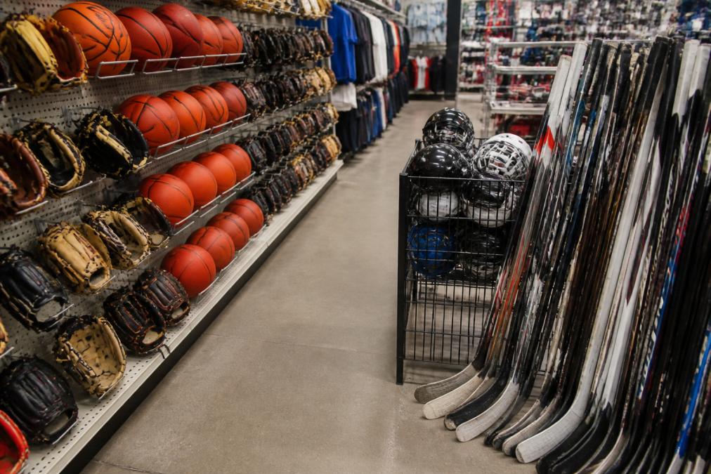 A clean retail aisle with new basketballs and baseball gloves on pegboard displays on one side, contrasting with a rack of clearly pre-owned hockey sticks and helmets on the other side of the store