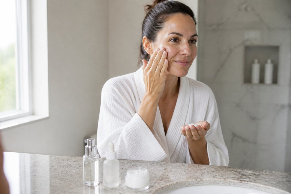 Woman applying skincare products in sequence on her face, showing hands with serum, moisturizer, and cream in a bright bathroom setting