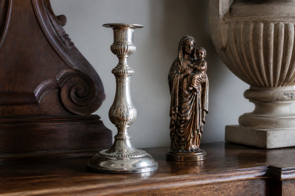 Close-up van een antiek zilveren kandelaar naast een koperen heiligenbeeldje op een donker houten dressoir, zacht daglicht dat de patina en glans benadrukt