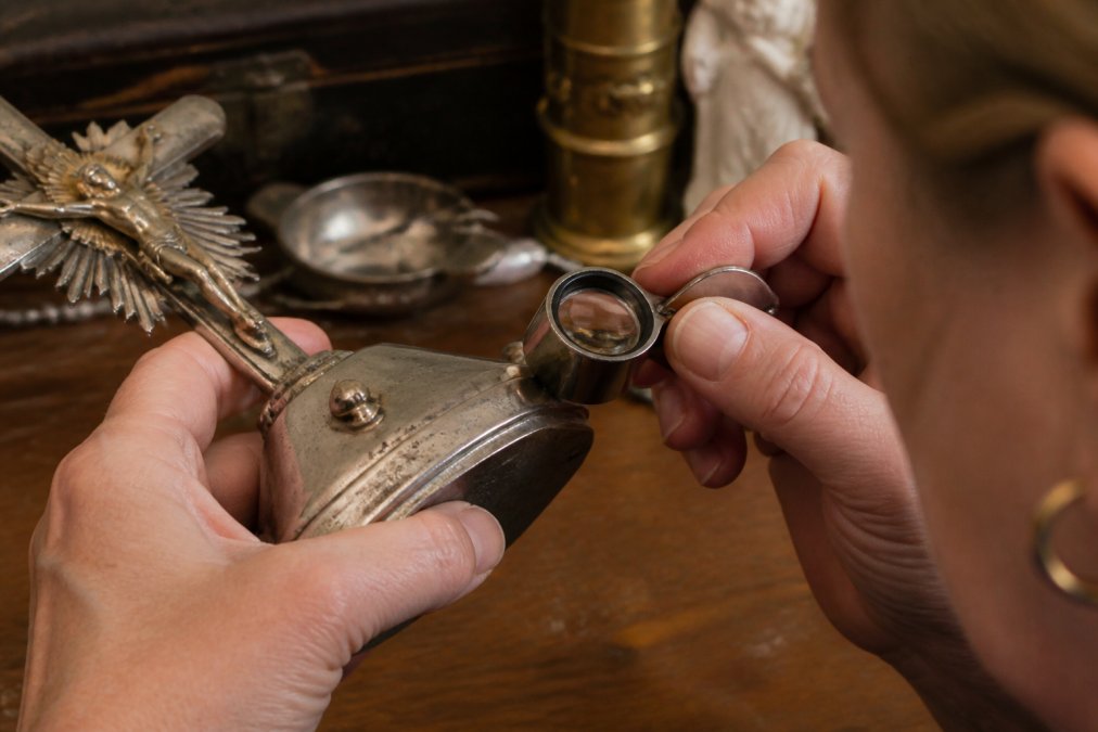 Iemand onderzoekt met een loep de stempels van antiek zilver of verzilverd devotionalia voor echtheidscontrole.