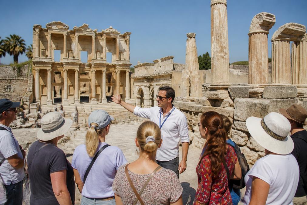 Profesyonel rehberin antik kent kalıntıları önünde turist grubuna tarihi yapıları gösterip anlatırken, taş sütunlar ve arkeolojik yapılar görülüyor, güneşli bir gün