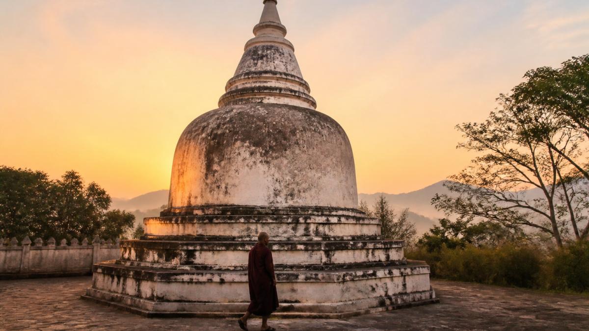 Ein verwitterter Stupa aus Stein in einem ruhigen Innenhof im Morgengrauen, dessen Kuppel sanftes orangefarbenes Licht einfängt, während eine einsame Gestalt im Uhrzeigersinn um seinen Sockel geht