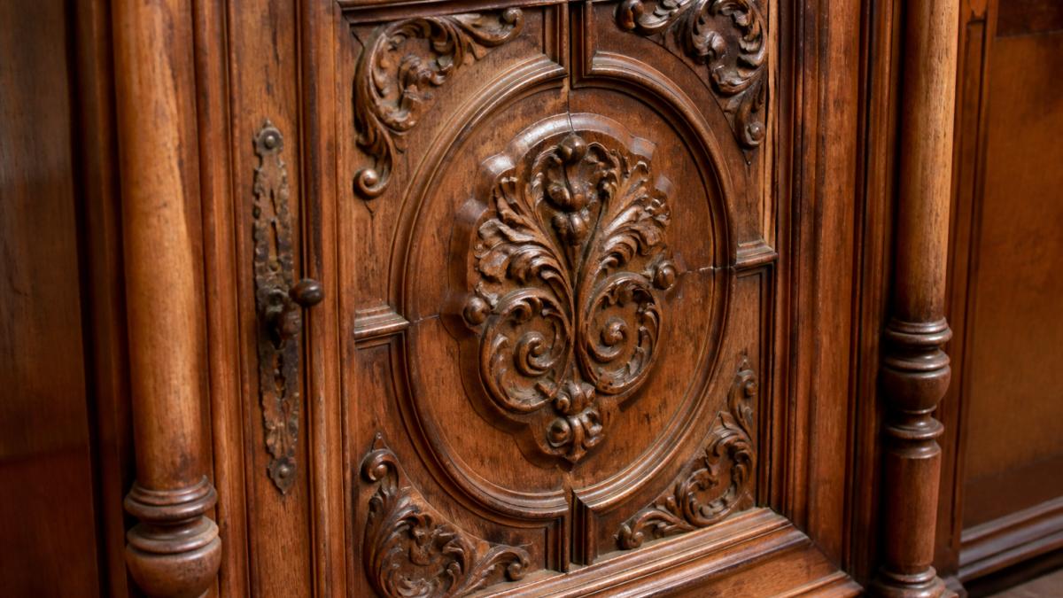 Close-up of ornately carved antique wooden cabinet door showing detailed hand-carved motifs, aged wood patina with visible grain variations, and aged finish in warm natural lighting