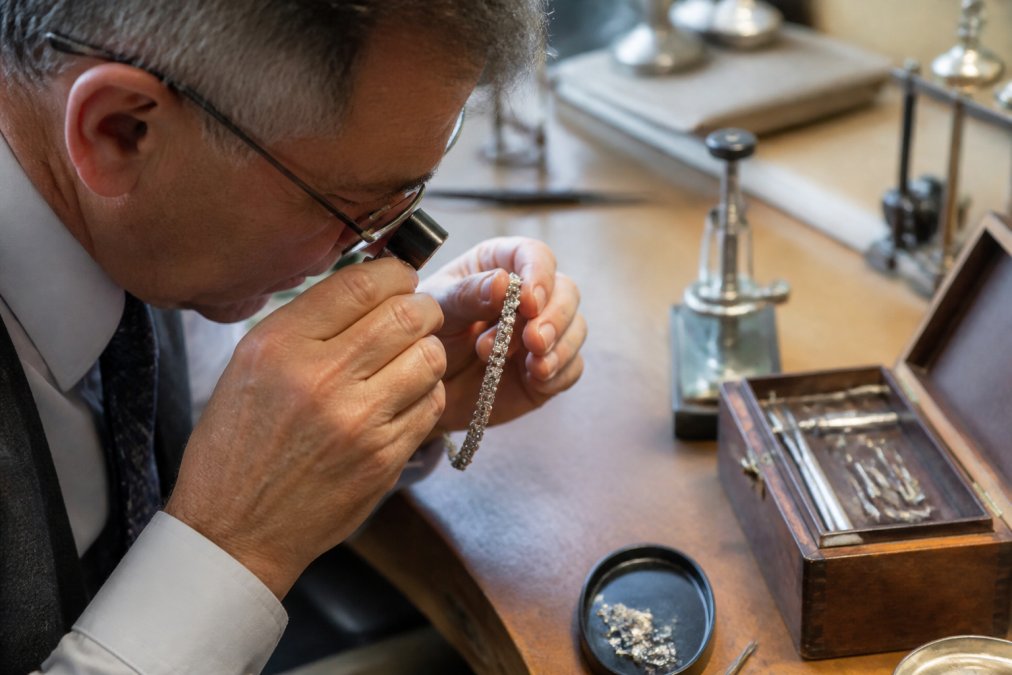 Professional jeweller examining antique diamond bracelet with loupe, viewing hallmarks and maker's marks clearly visible on platinum setting under natural workshop lighting