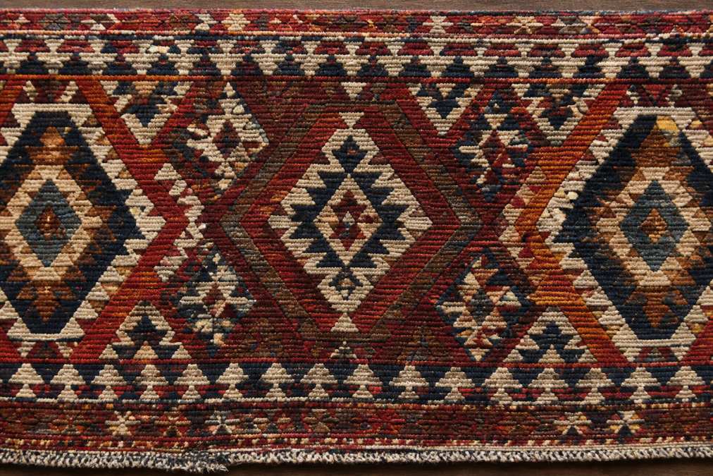 Close-up overhead shot of an antique kilim rug displaying intricate geometric patterns and tribal motifs in rich burgundy, navy, and cream vegetable dye colors, with natural daylight highlighting the flat-weave texture and pattern intricacy