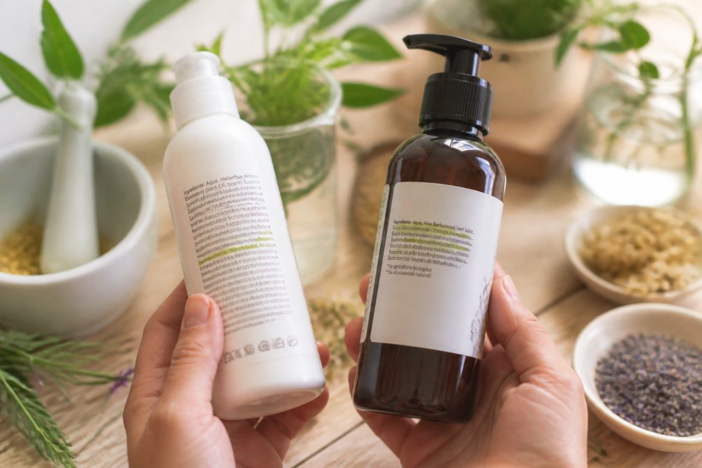Mani che tengono due flaconi di cosmetici naturali con etichette INCI ben visibili su un piano in legno con piante aromatiche