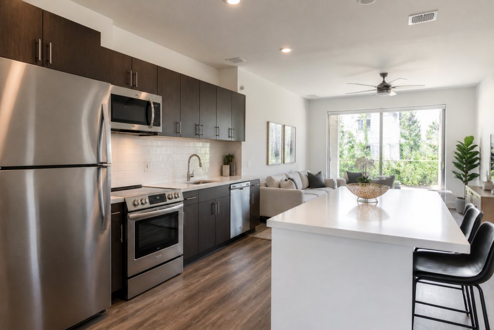 Modern apartment interior with stainless steel appliances, quartz countertops, bright natural lighting from large windows, and contemporary furnishings
