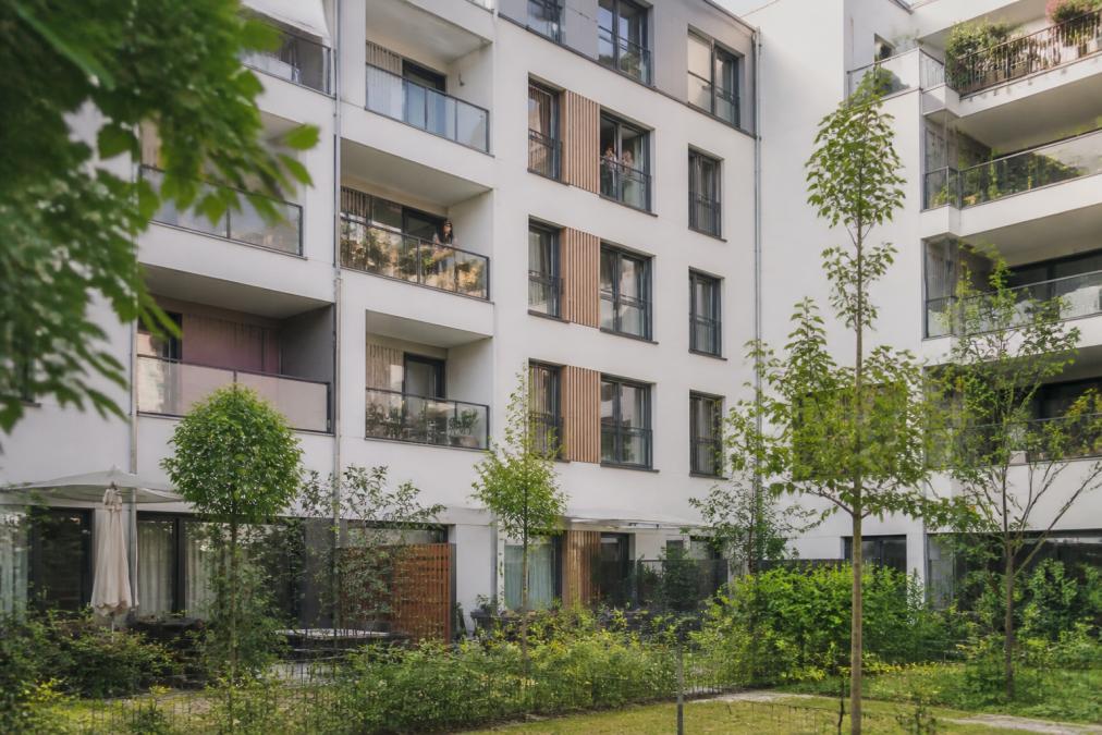 Vue d'une façade d'immeuble résidentiel moderne avec balcons individuels, jardins partagés en rez-de-chaussée, et habitants visibles aux fenêtres dans un quartier urbain densifié