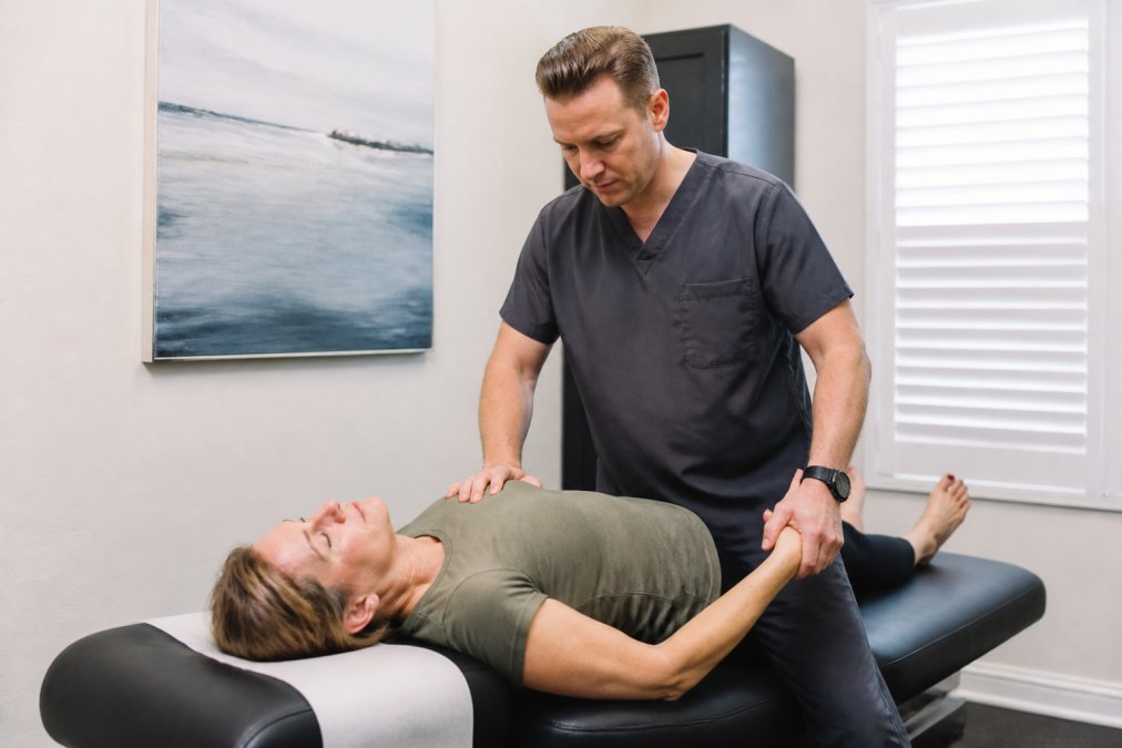 Patient lying on treatment table while practitioner performs spinal assessment and extremity evaluation, calm clinical environment with soft lighting and professional medical setting