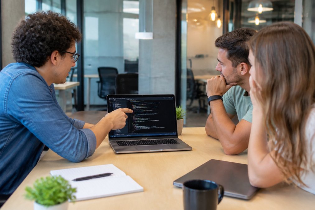 Person presenting a data science project on a laptop screen to two colleagues in a casual modern office setting, with code and visualizations visible on the screen under warm office lighting