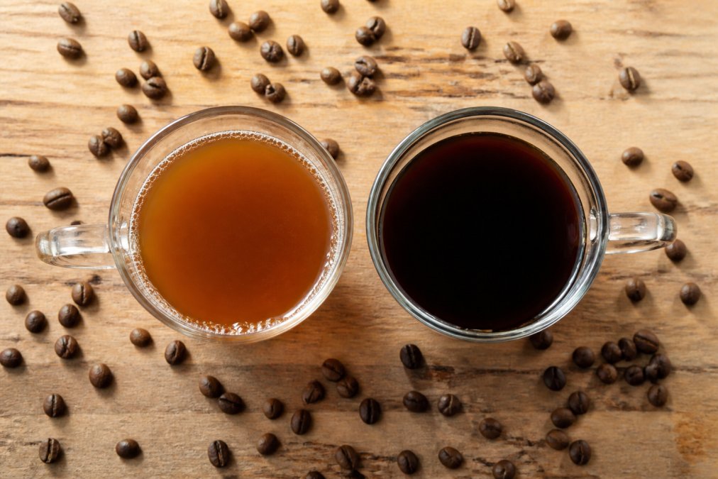 A close-up overhead shot of two identical glass cups of black coffee side by side on a light wood table, one filled with a lighter golden-brown brew and the other with a darker richer-looking brew, whole roasted coffee beans scattered around the cups