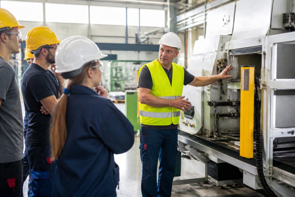 Ein Schulungsleiter in Warnweste demonstriert einer kleinen Gruppe von Mitarbeitern mit Schutzhelmen die Sicherheitsvorrichtungen an einer Produktionsmaschine, Halle mit natürlichem Lichteinfall