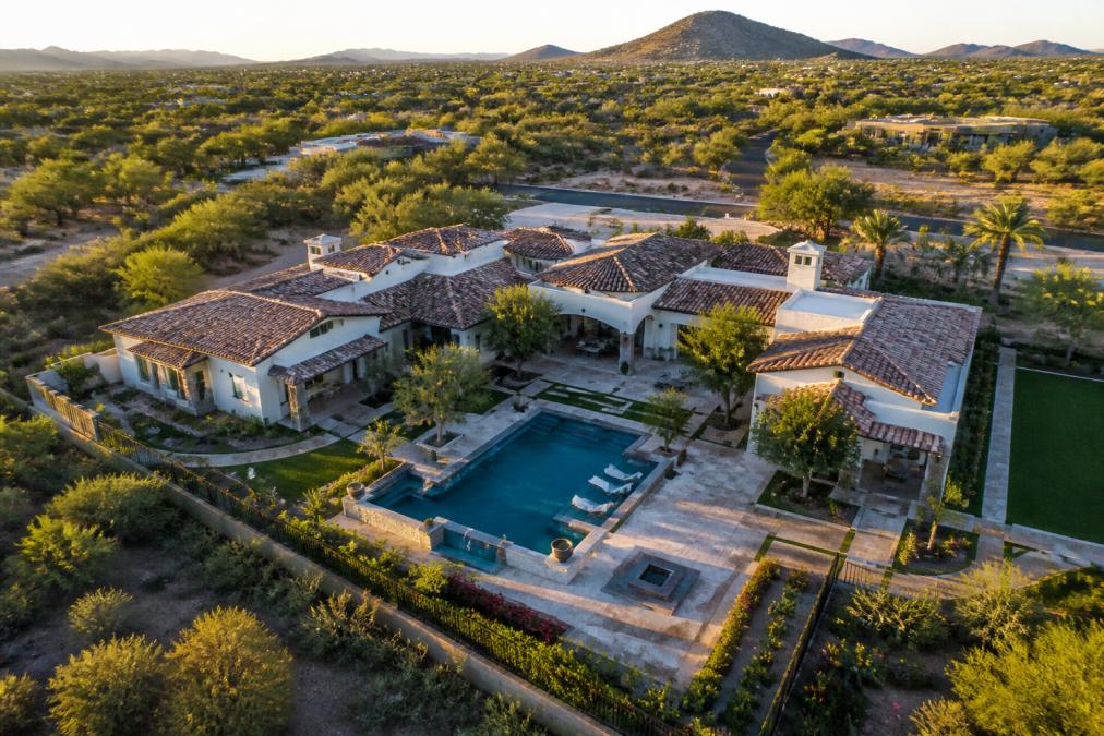 Aerial view of a luxury estate captured from drone perspective showing the complete architectural roofline, resort-style pool with surrounding pavers, manicured gardens with mature trees, and the property's relationship to surrounding landscape in late afternoon golden light