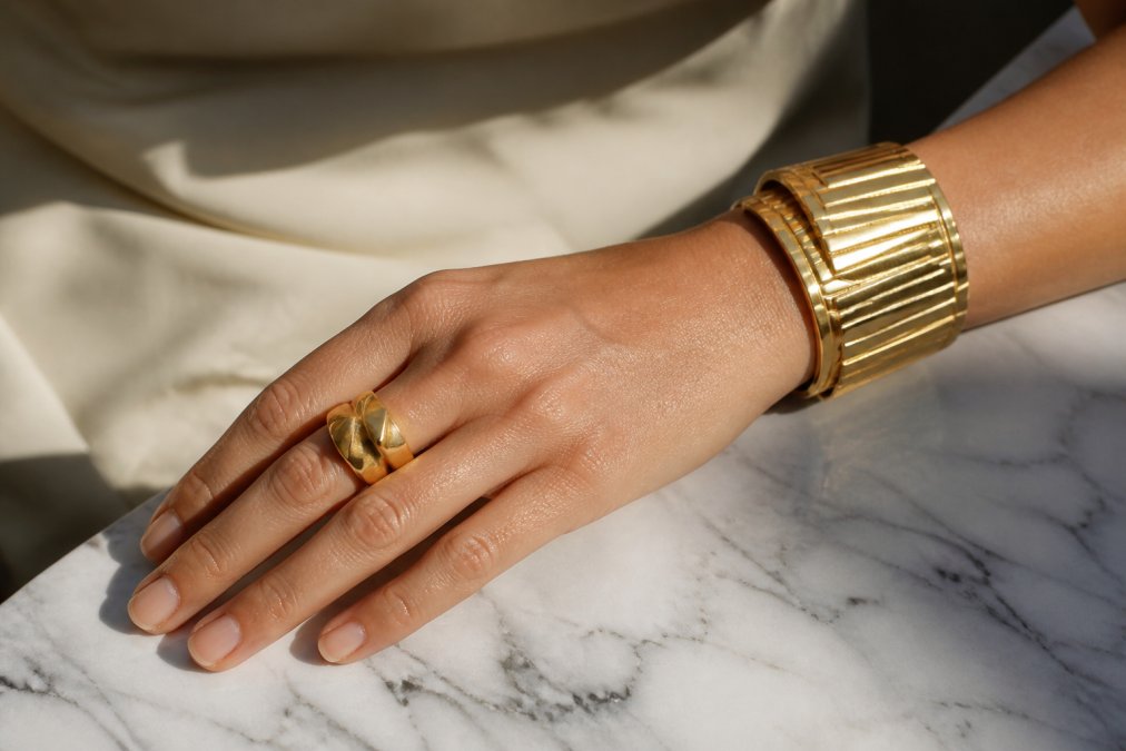 Close-up photograph of a woman's hand wearing a thick sculptural gold ring and a heavy gold cuff bracelet, resting on a white marble surface, soft directional daylight catching the geometric textures