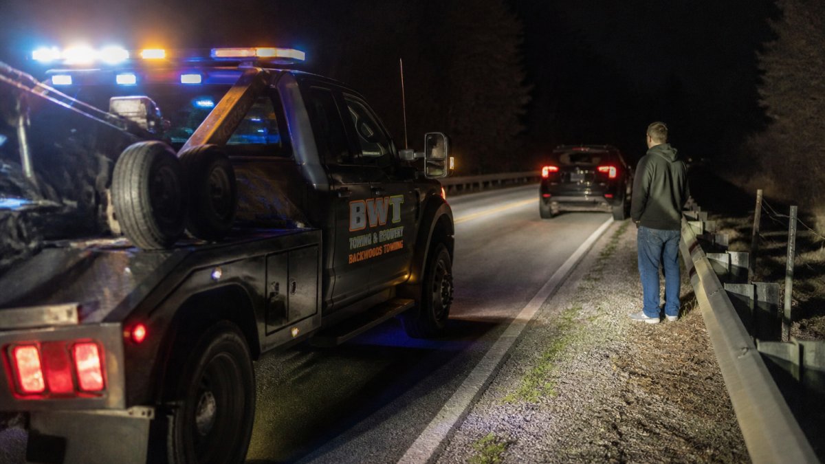Tow truck with emergency lights activated at night on a rural road, disabled vehicle ahead with driver standing safely behind guardrail