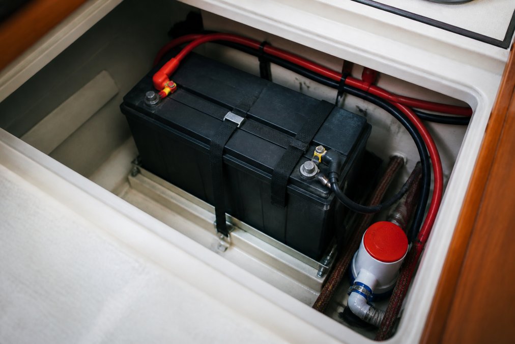 Close-up of a sealed AGM marine battery strapped into a sailboat's fiberglass battery compartment, with marine-grade wiring and a bilge pump visible in the dim cabin light