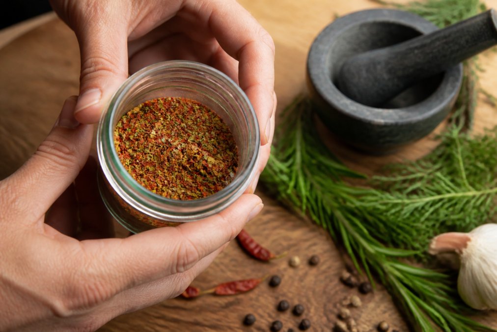 Close-up of hands holding a small glass jar of colorful spice blend over a wooden cutting board with fresh herbs and scattered whole spices nearby, warm natural kitchen lighting