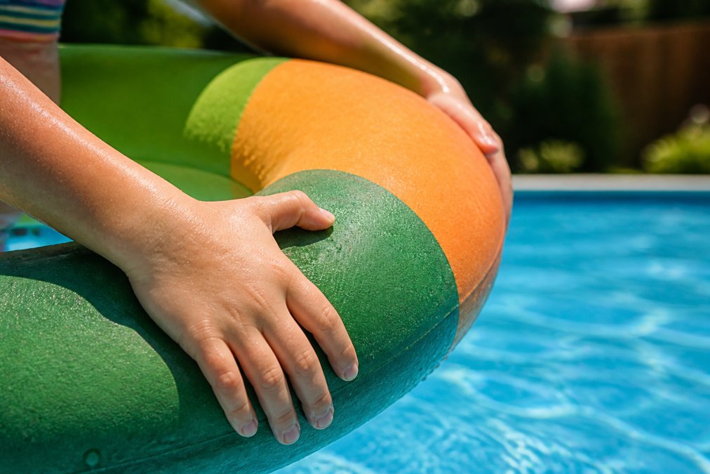 Close-up of a child's hands gripping a vibrant biodegradable pool float in a backyard swimming pool under bright afternoon sunlight, showing the toy's textured natural rubber surface and colorful design