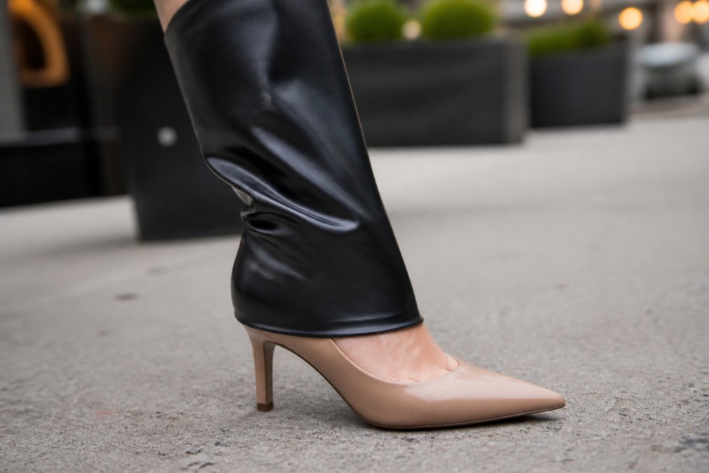Close-up of a woman's leg showing a black stretch faux leather boot sleeve pulled over a nude pump heel, demonstrating the seamless transition from shoe to sleeve