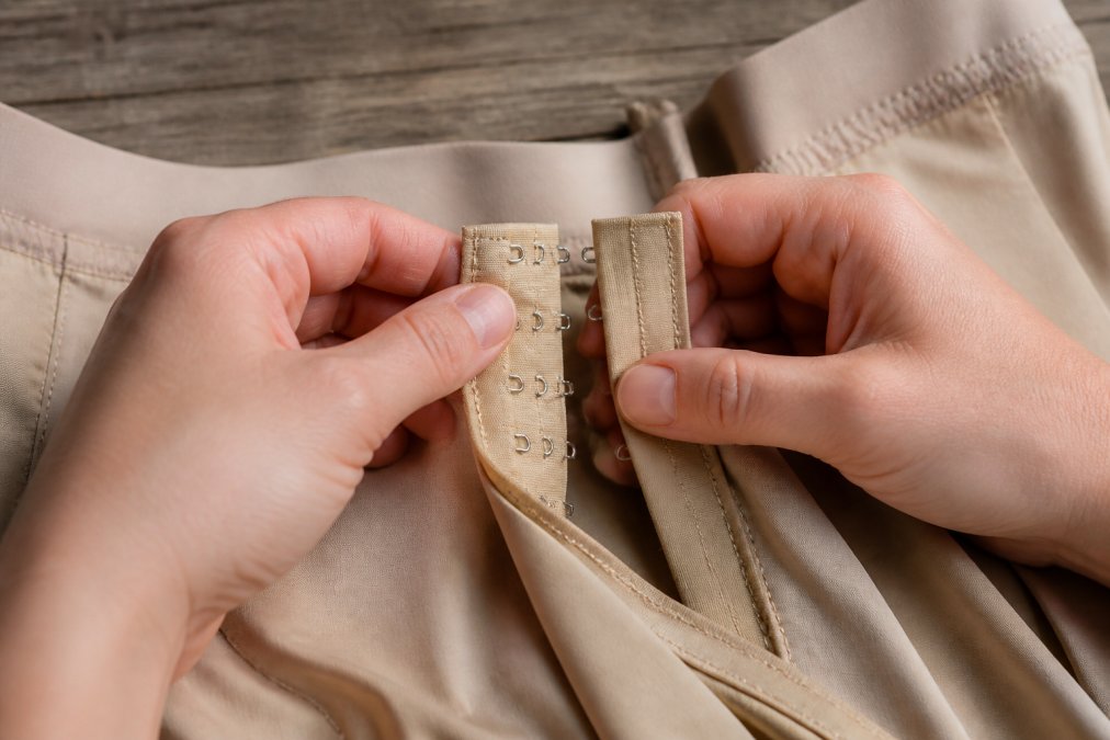 Close-up of hands carefully examining and adjusting a hook-and-eye closure on a beige shapewear piece, showing the hardware detail and proper handling technique