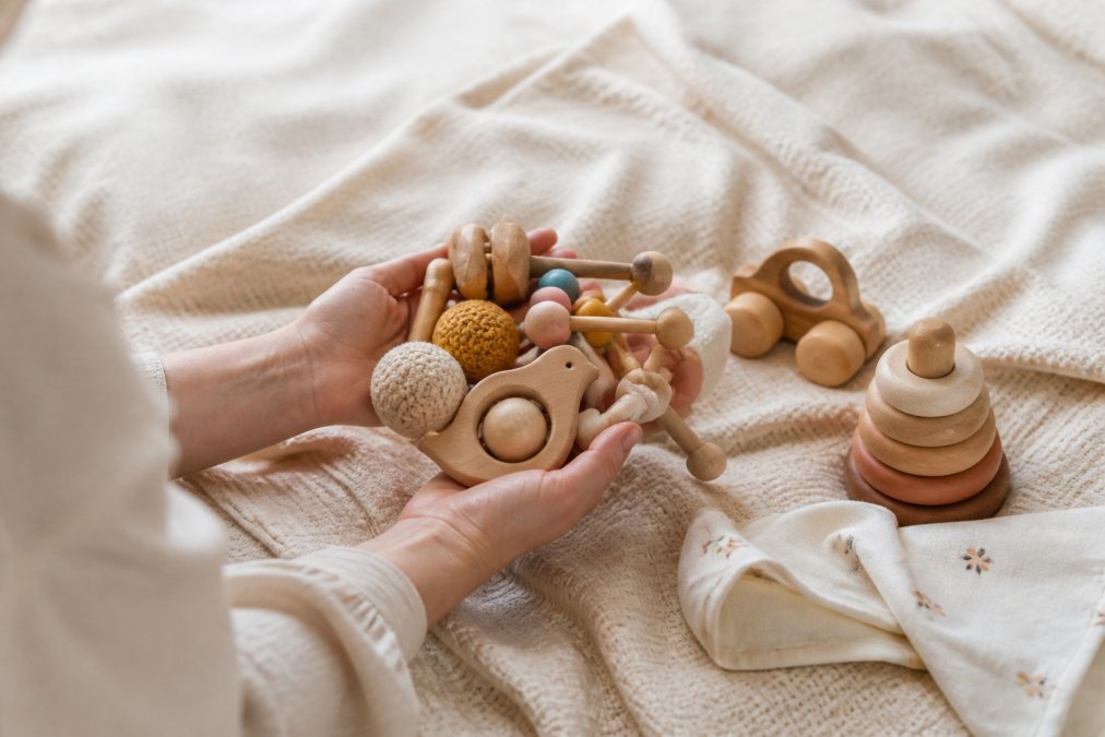 A parent's hands holding a selection of wooden and sensory baby toys arranged on a soft neutral blanket in warm natural light