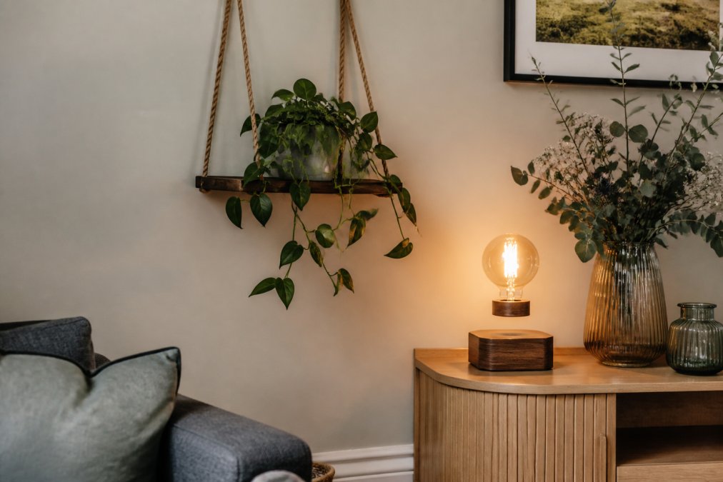 A styled living room corner in a British home showing a wooden hanging swing shelf holding a trailing plant, with a magnetic levitation floating bulb lamp glowing on a side table nearby
