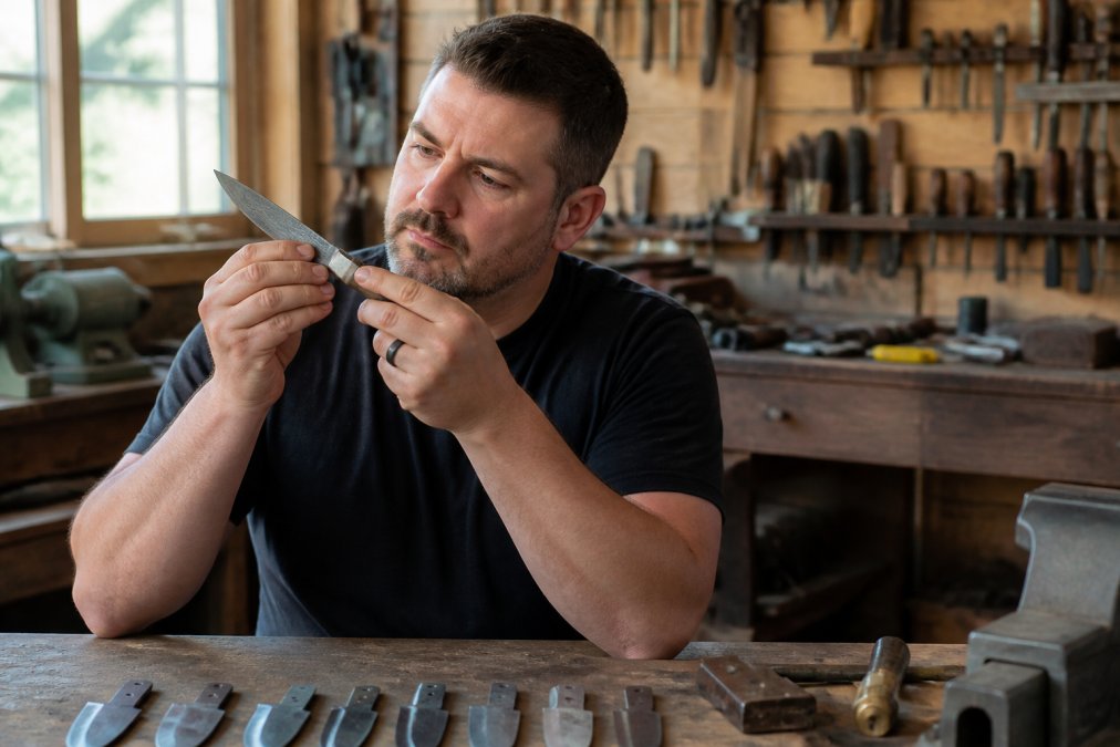 Custom knife maker inspecting a finished blade for sharpness in a workshop filled with steel blanks and tools