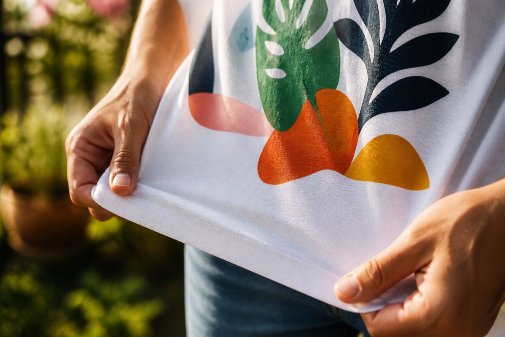 Close-up of hands holding the hem of a soft cotton t-shirt, showing the fabric texture and a vibrant custom print design in natural sunlight