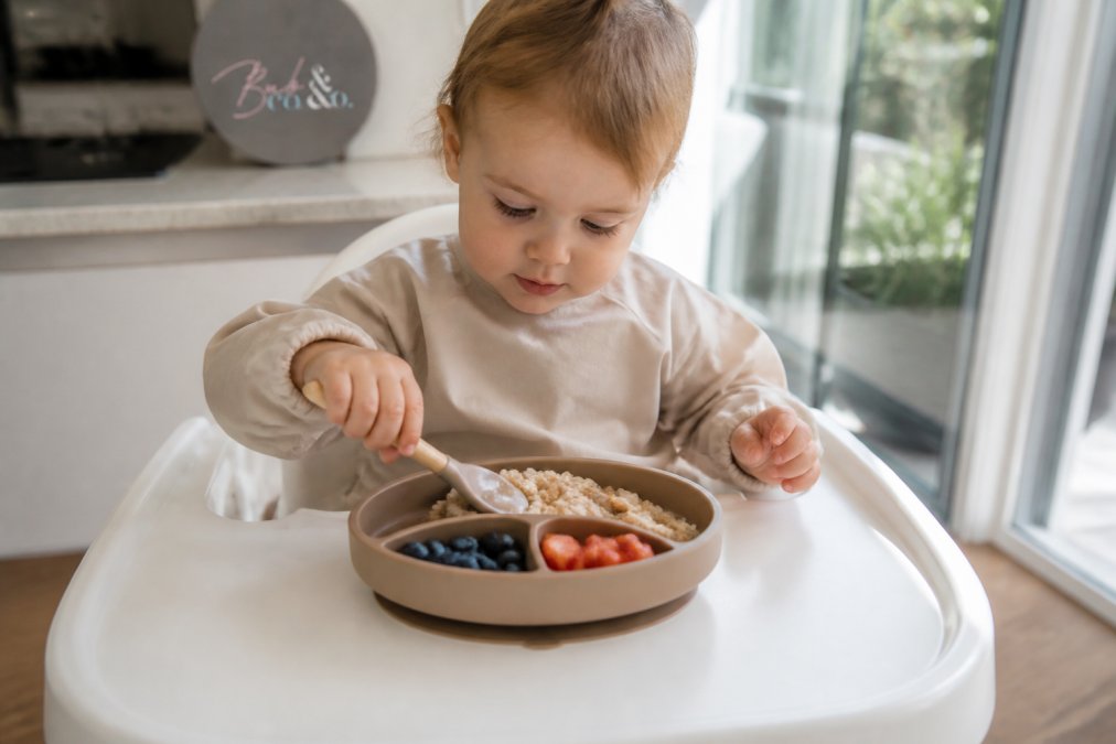Toddler learning to self-feed using a silicone suction divided plate on a highchair tray in a sunny kitchen