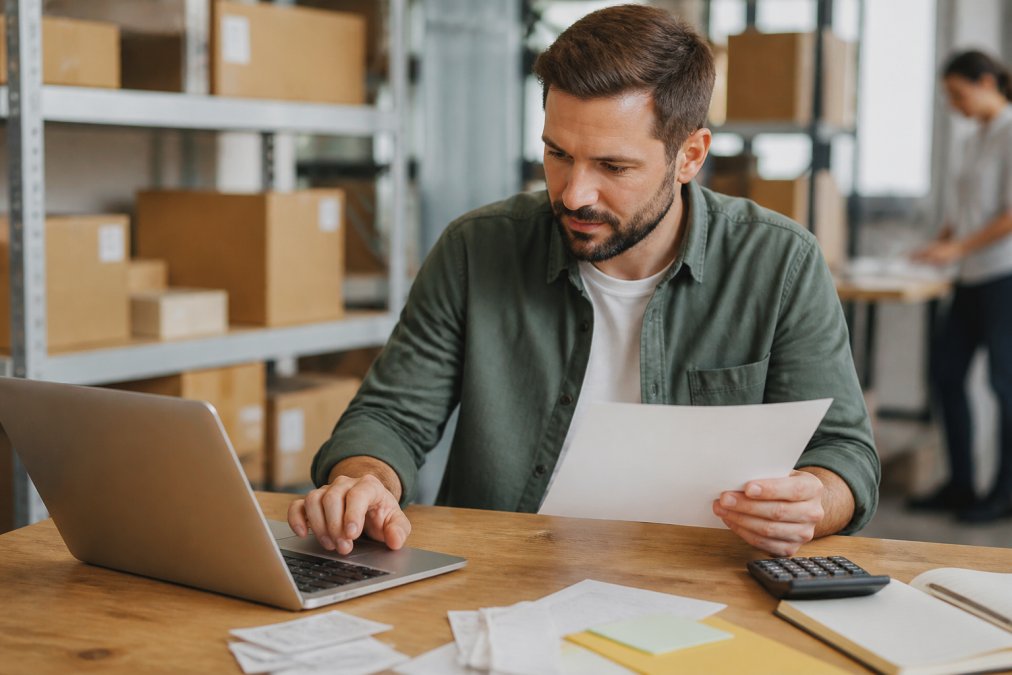 Business owner reviewing fulfillment metrics and cost spreadsheets on a laptop at a desk, with shipping labels, inventory notes, and a calculator visible, natural office lighting