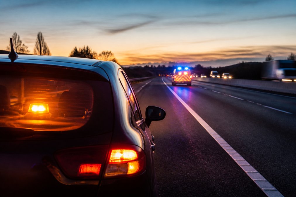 Are Emergency Beacons Worth the Investment? Breakdown scenario on a busy motorway at dusk: a stationary vehicle with hazard lights activated, high-visibility beacon flashing on the dashboard, and an emergency services vehicle approaching in the distance