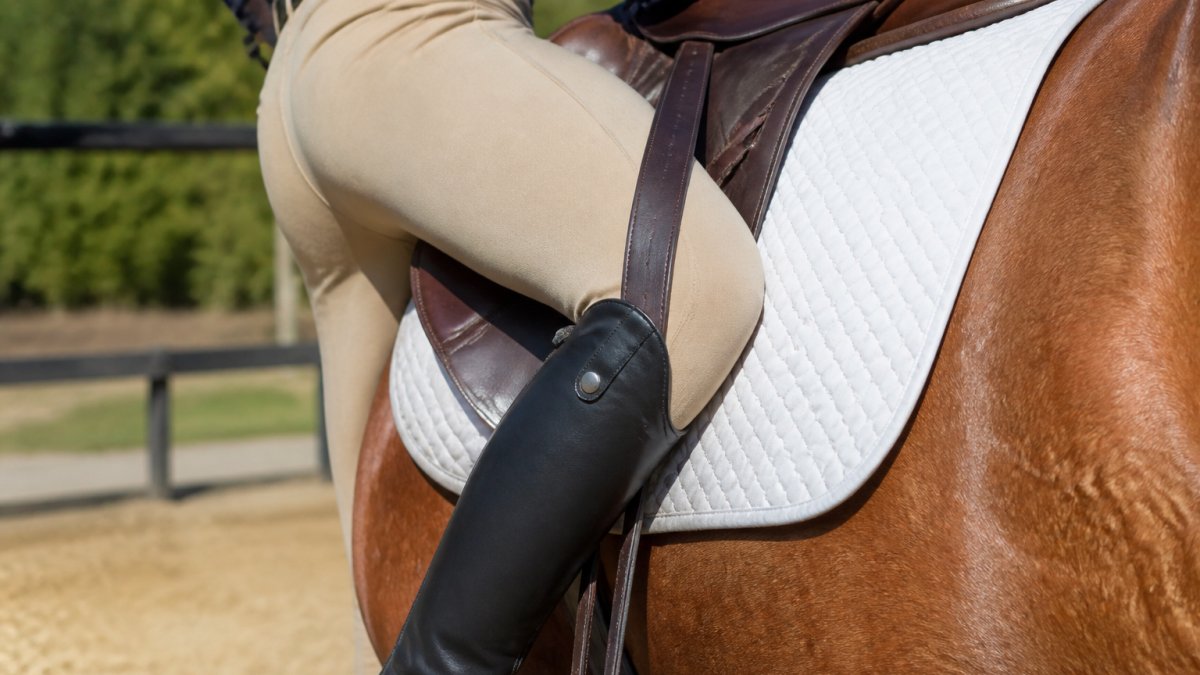 Close-up of a rider's leg positioned against a saddle during mounting, showing proper contact between the wide leather and the rider's calf and lower leg
