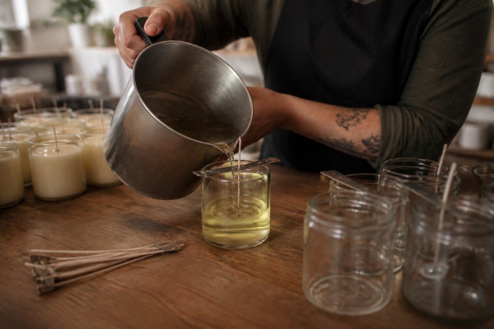 Close-up of a craftsperson's hands carefully pouring melted wax into a clear glass jar on a wooden workbench, warm workshop lighting, small batches in the background
