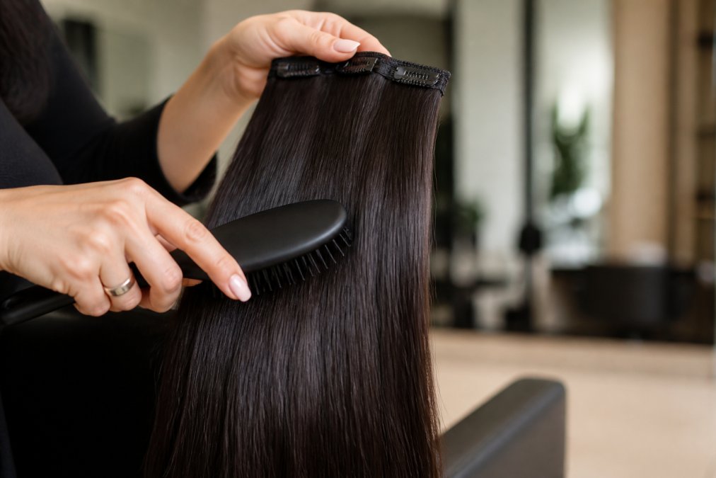 Close-up of a hairstylist's hands gently brushing long, glossy natural-looking hair extensions, with a soft salon background