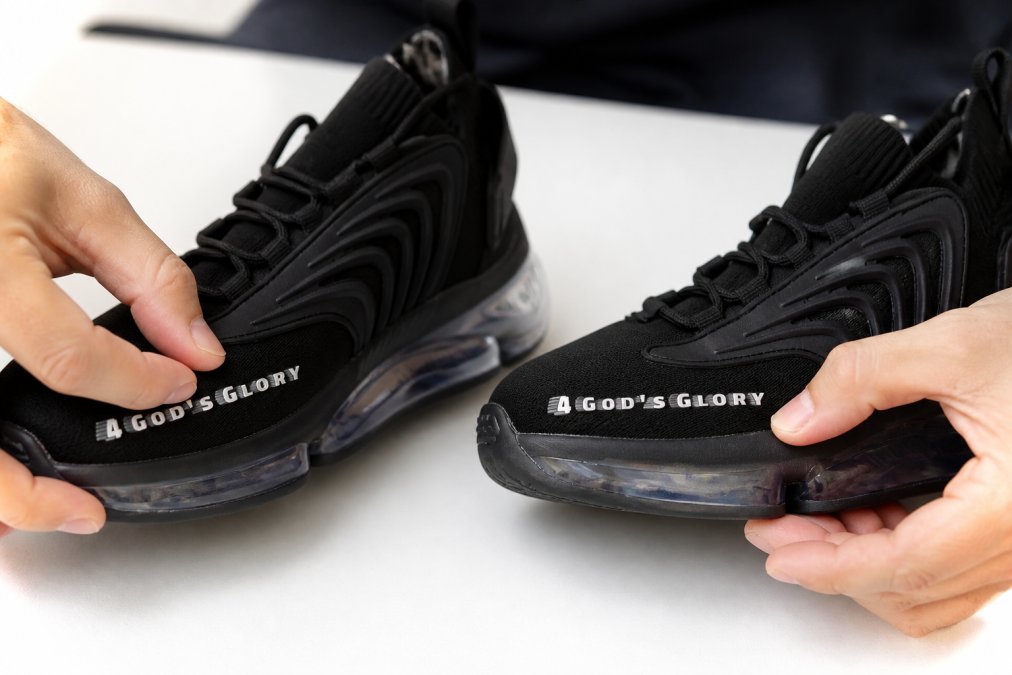 Close-up of hands examining sneaker stitching details, comparing authentic Nike Air Max logo embroidery with counterfeit version on white work surface with natural lighting
