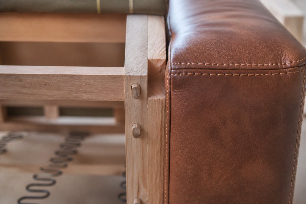 Close-up of premium sofa frame showing kiln-dried hardwood joints with visible dowel construction, hand-stitched upholstery seams with tight, even stitching, and exposed wooden frame details
