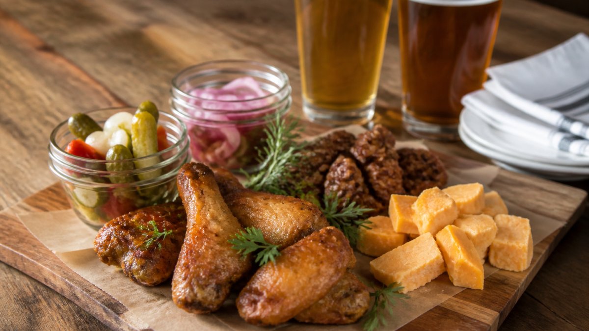 Close-up of a spread of premium bar snacks on a wooden board: crispy wings with golden skin, aged cheddar chunks, candied walnuts, pickled vegetables in small glass jars, and fresh herb garnishes, with beer glasses and napkins in the background on a rustic table