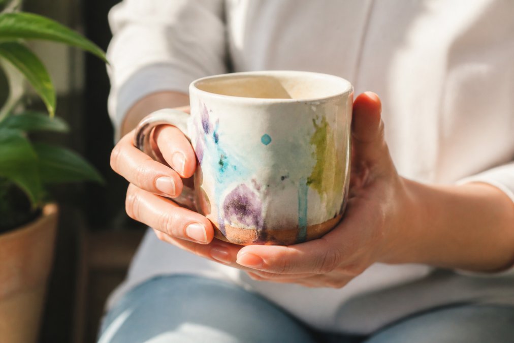 Close-up of hands cradling a ceramic handmade mug with watercolor artwork, warm natural sunlight illuminating the artisanal glaze details and brushwork