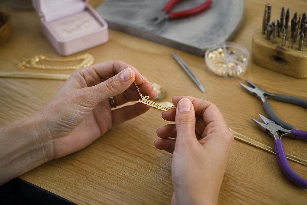 Close-up of hands holding a delicate personalized name necklace with a cursive pendant, soft natural light, jewelry box in the background