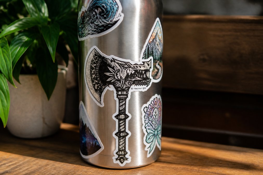 Close-up of a stainless steel water bottle decorated with multiple detailed handmade vinyl stickers showing intricate line work, vibrant colors, and die-cut edges, sitting on a wooden table in natural sunlight