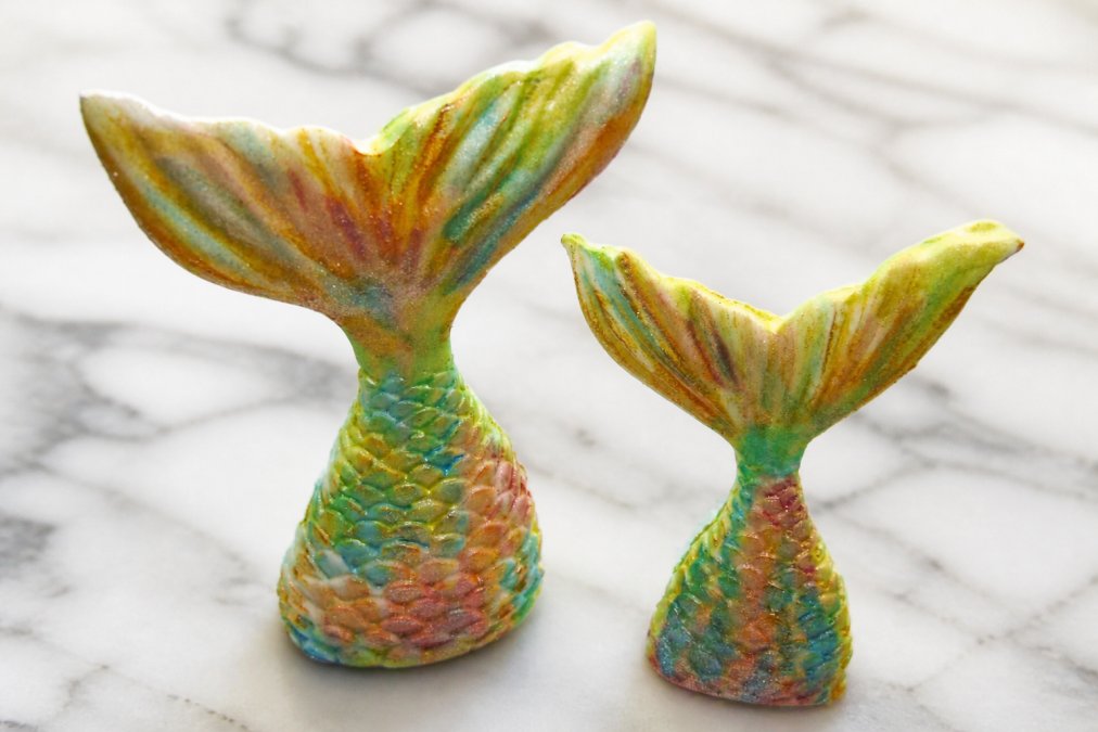Close-up of colourful handmade sugar paste cake toppers with edible glitter finish arranged on a white marble surface under soft natural light, showing fine hand-sculpted detail