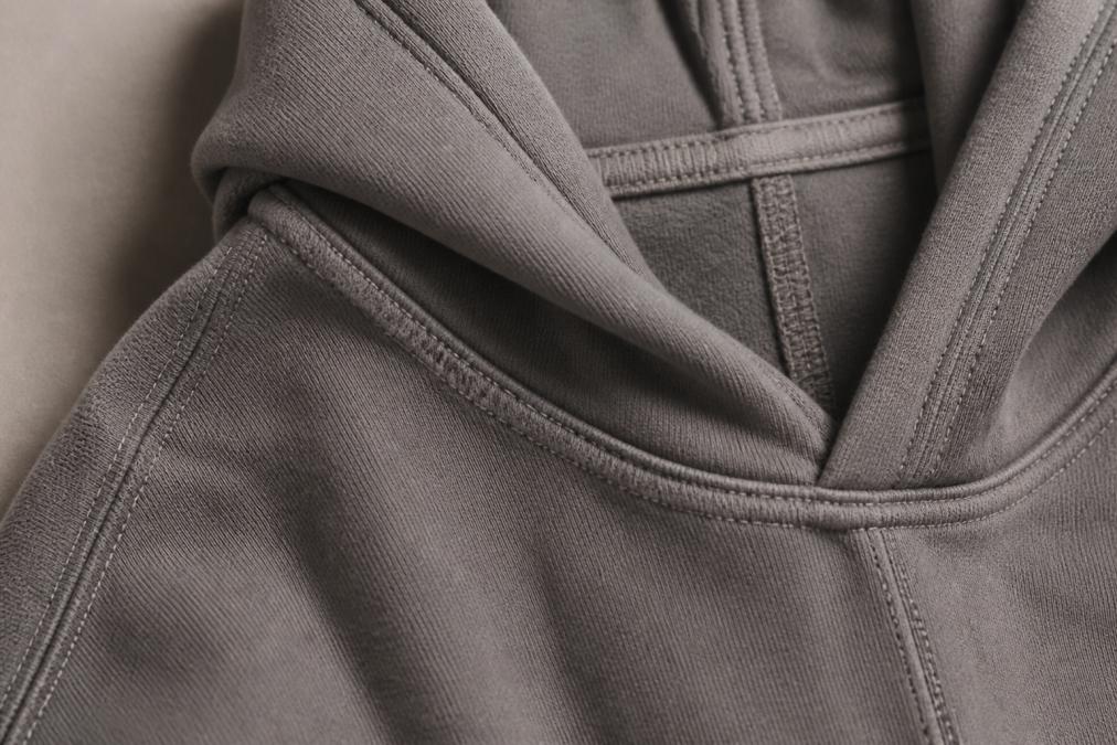 Close-up detail of a heavyweight hoodie's double-stitched side seams, flatlock hood construction with reinforced stitching, and binding-taped neckline showing dense fabric texture and premium seam craftsmanship