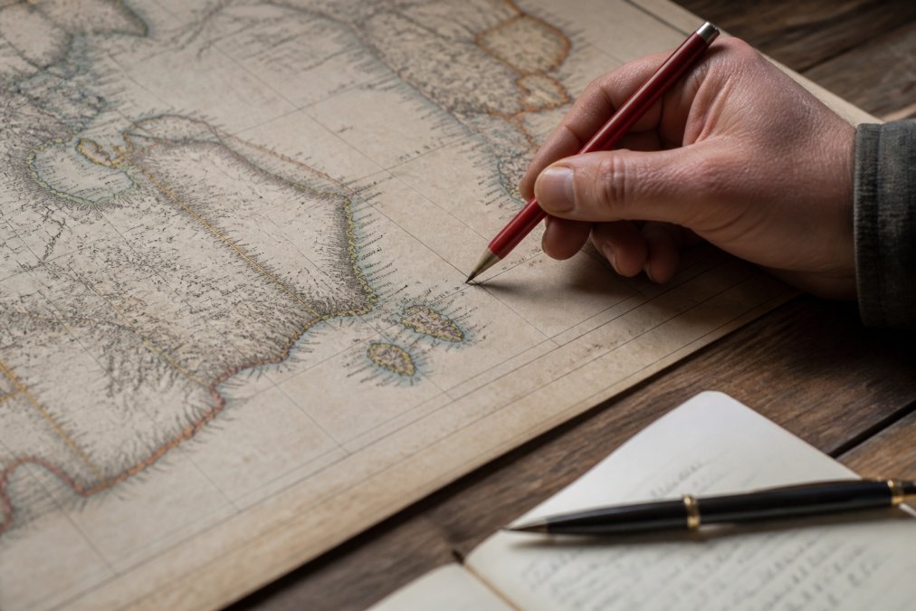 A close-up photograph of a printed vintage map spread on a wooden table, with a hand holding a red pen hovering over a coastline, suggesting someone annotating or correcting place names
