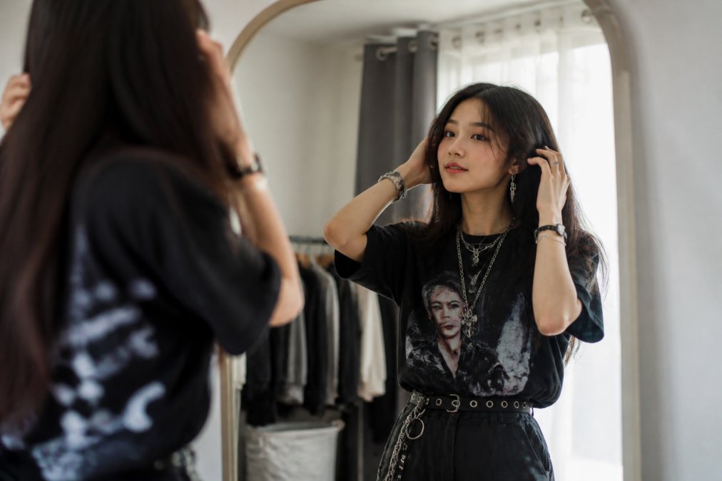 Young person standing in front of a mirror in natural light, wearing a custom-printed K-pop streetwear outfit, confidently styling themselves with accessories and considering their personal aesthetic