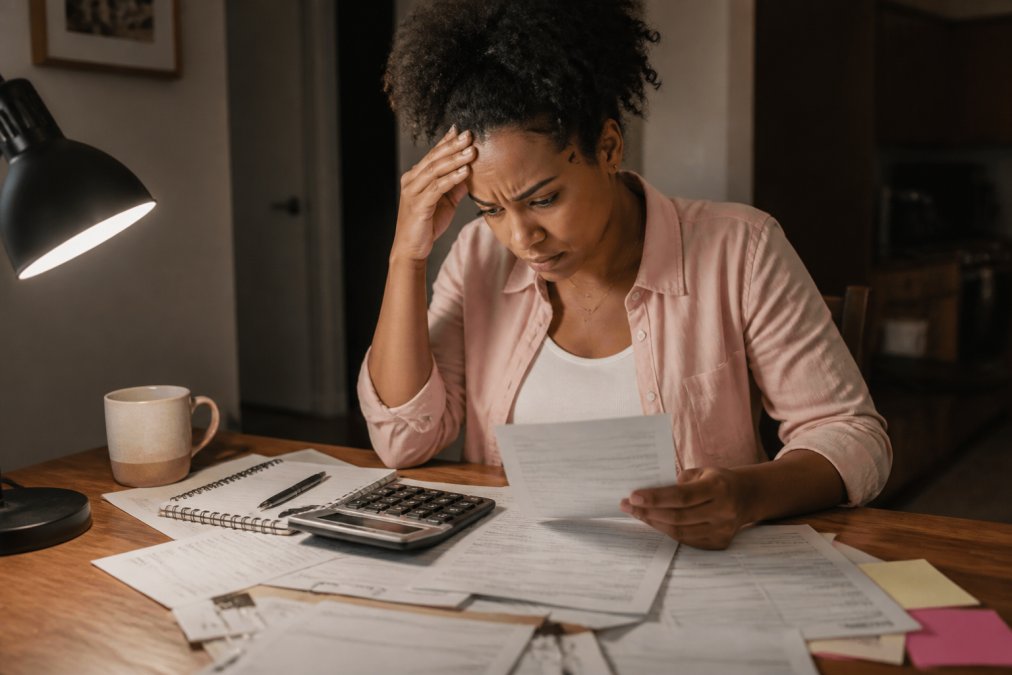 Person sitting at a desk reviewing bills and payment statements with a concerned expression, calculator and notepad visible on wooden desk under warm lamp lighting