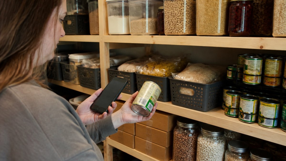 A parent in a home pantry scanning a can's barcode with a smartphone while shelves of labeled storage containers and date-marked boxes stand behind them