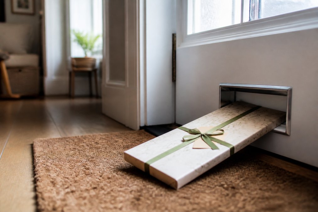 A slim, neatly wrapped letterbox gift box resting on a doormat just inside a front door, partially through the letterbox slot, with soft natural light filtering through a hallway window