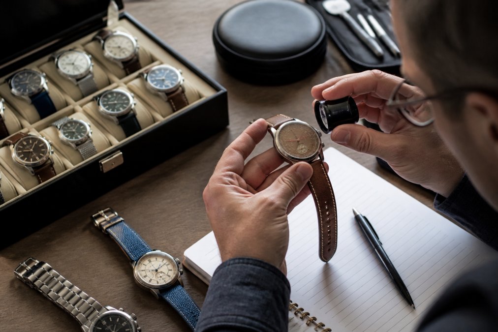 Watch collector examining a timepiece at a desk with reference materials, valuation notes, and a loupe under task lighting, with multiple watches displayed on the workspace