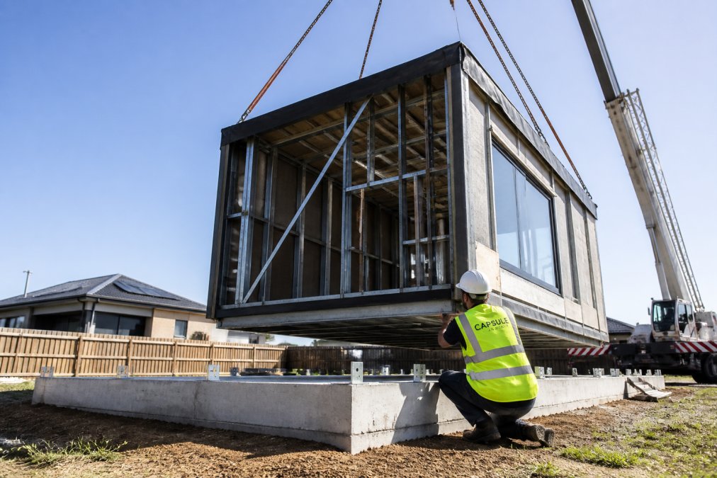 A steel-framed modular home module being craned onto a permanent concrete footing on a suburban block, with a building surveyor in high-visibility vest inspecting the sub-floor connection